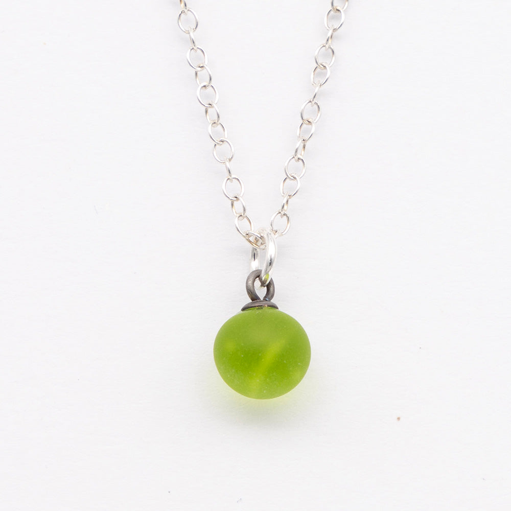 Close up of a lime green frosted glass ball charm on a plain white background. The charm is frosted. It hangs on a small silver jump ring. The necklace is completed with a silver chain.