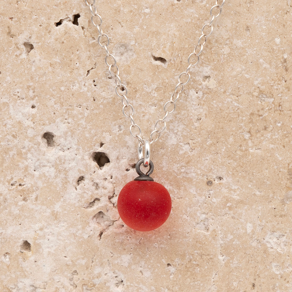 Close up of a orange frosted glass ball charm on a sandstone background. The charm is frosted. It hangs on a small silver jump ring. The necklace is completed with a silver chain.