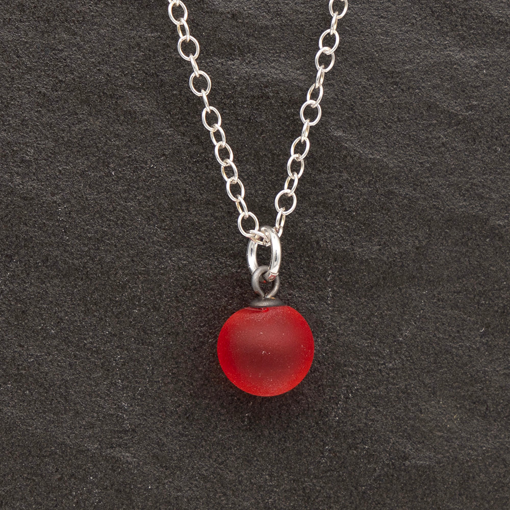 Close up of an orange frosted glass ball charm on a dark slate background. The charm is frosted. It hangs on a small silver jump ring. The necklace is completed with a silver chain.