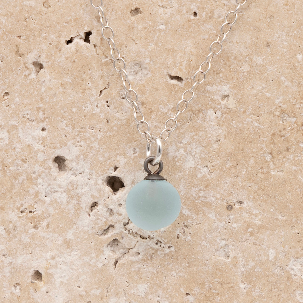 Close up of apale blue frosted glass ball charm on a sandstone background. The charm is frosted. It hangs on a small silver jump ring. The necklace is completed with a silver chain.