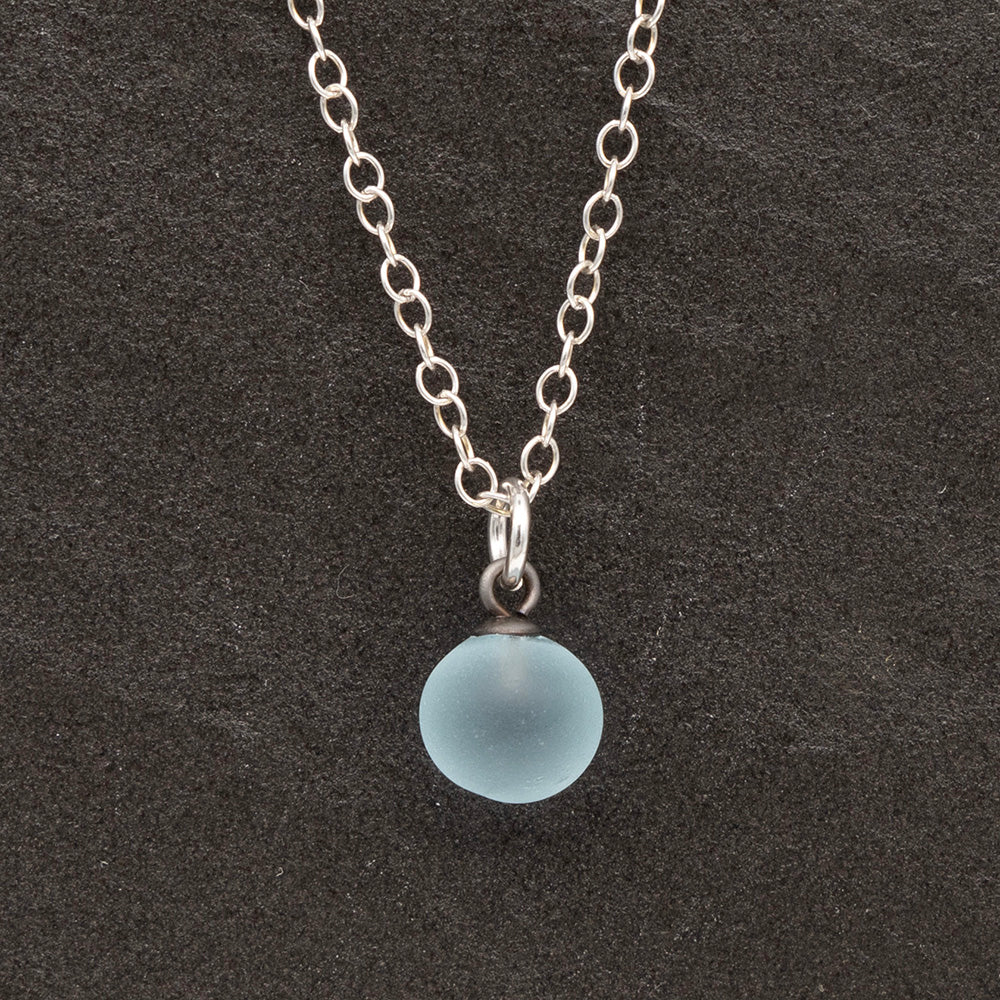 Close up of a pale blue frosted glass ball charm on a dark slate background. The charm is frosted. It hangs on a small silver jump ring. The necklace is completed with a silver chain.