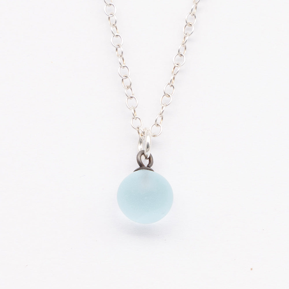 Close up of a pale blue frosted glass ball charm on a plain white background. The charm is frosted. It hangs on a small silver jump ring. The necklace is completed with a silver chain.