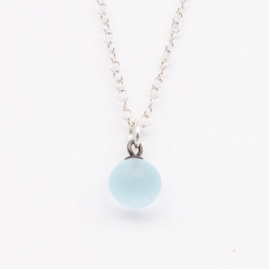 Close up of a pale blue frosted glass ball charm on a plain white background. The charm is frosted. It hangs on a small silver jump ring. The necklace is completed with a silver chain.