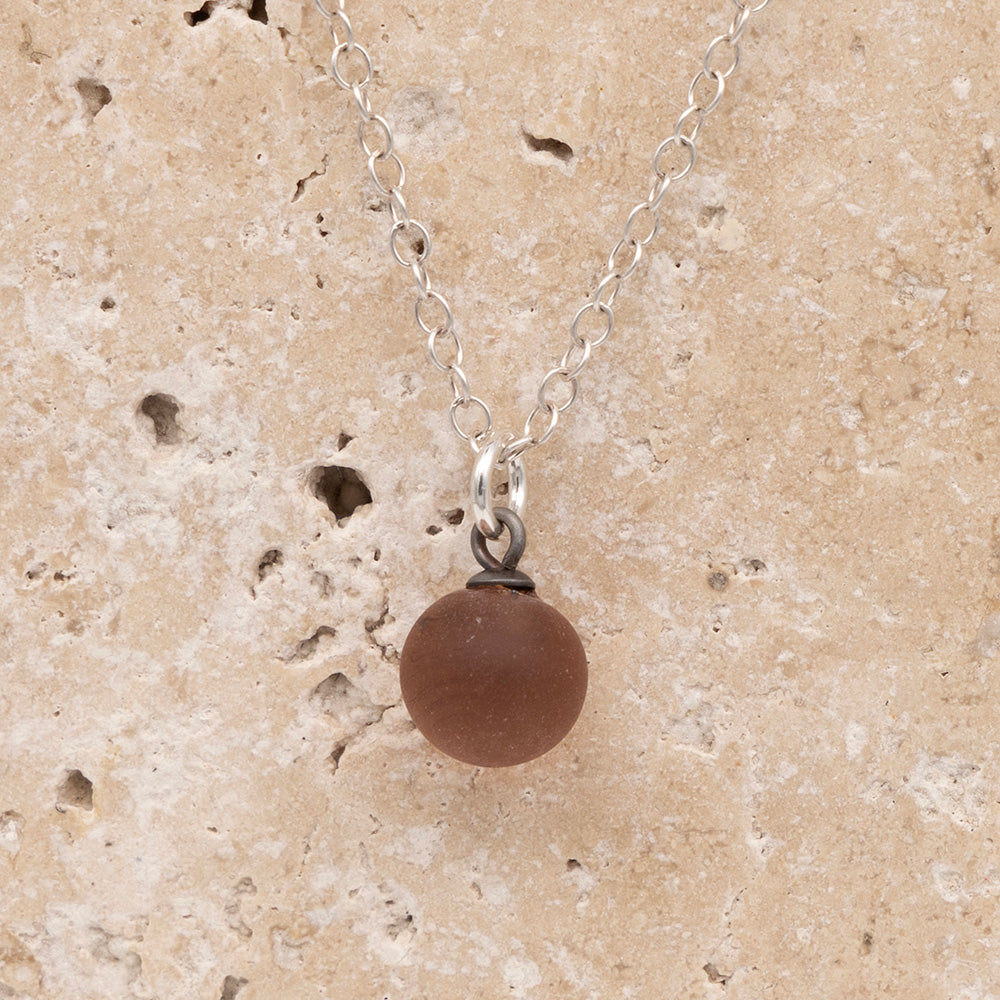 Close up of a pink frosted glass ball charm on a sandstone background. The charm is frosted. It hangs on a small silver jump ring. The necklace is completed with a silver chain.