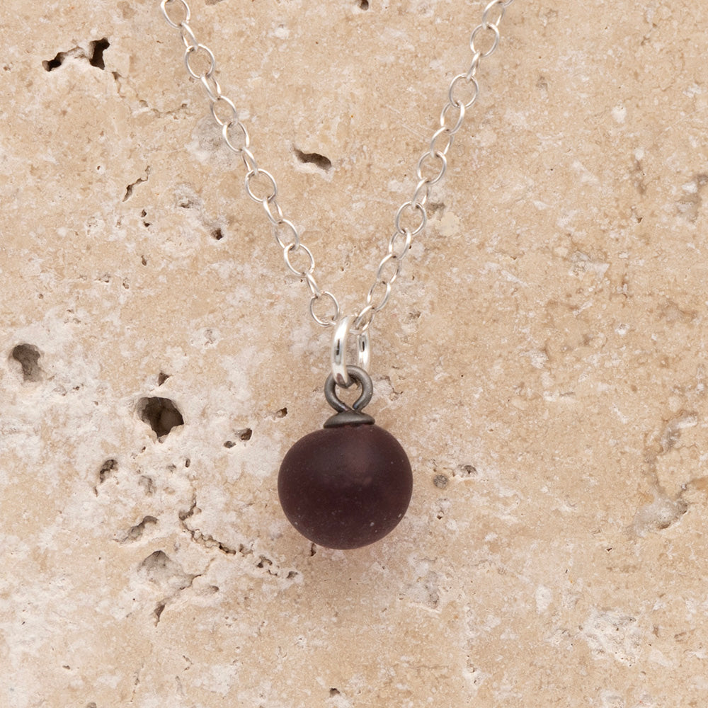 Close up of a purple frosted glass ball charm on a sandstone background. The charm is frosted. It hangs on a small silver jump ring. The necklace is completed with a silver chain.