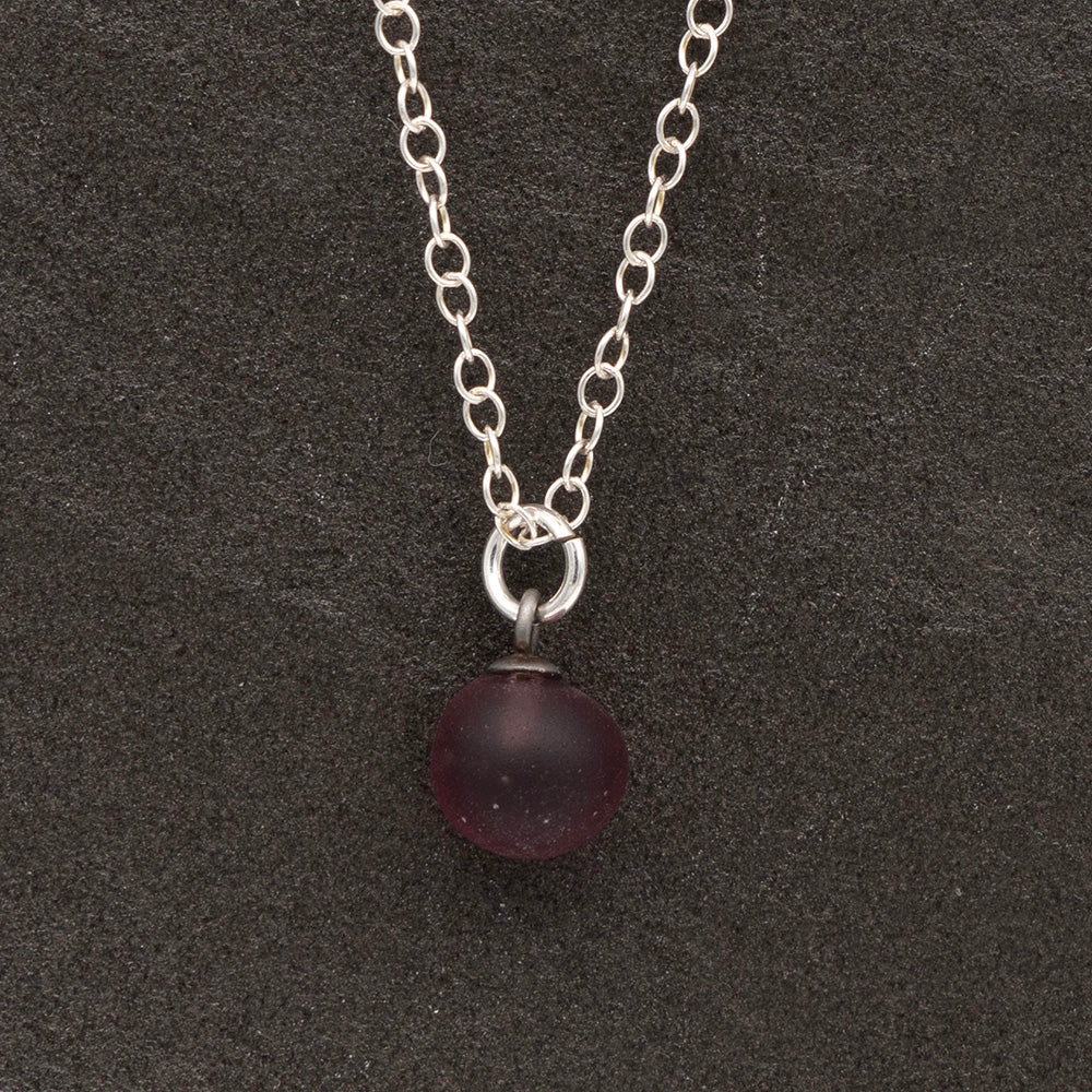 Close up of a purple frosted glass ball charm on a dark slate background. The charm is frosted. It hangs on a small silver jump ring. The necklace is completed with a silver chain.