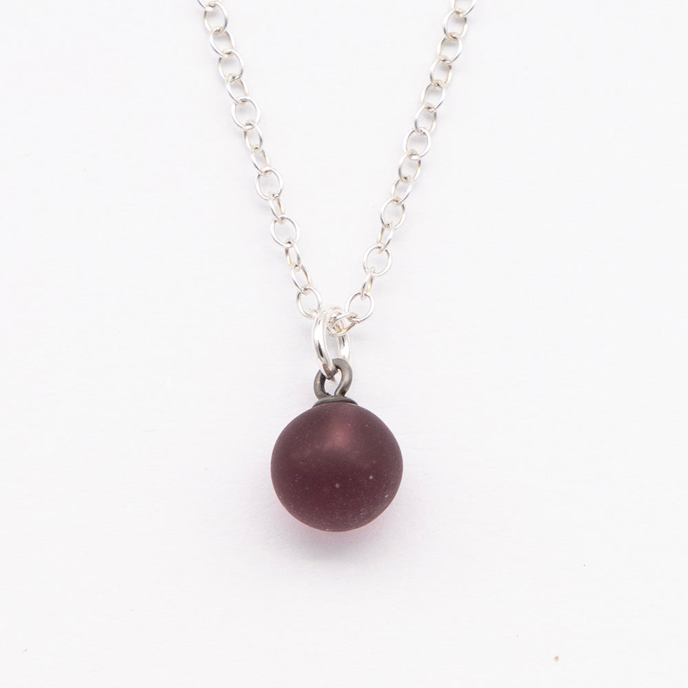 Close up of a purple frosted glass ball charm on a plain white background. The charm is frosted. It hangs on a small silver jump ring. The necklace is completed with a silver chain.