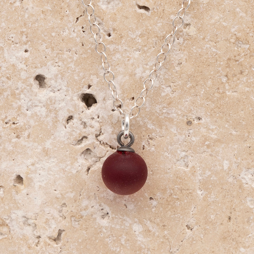 Close up of a red frosted glass ball charm on a sandstone background. The charm is frosted. It hangs on a small silver jump ring. The necklace is completed with a silver chain.