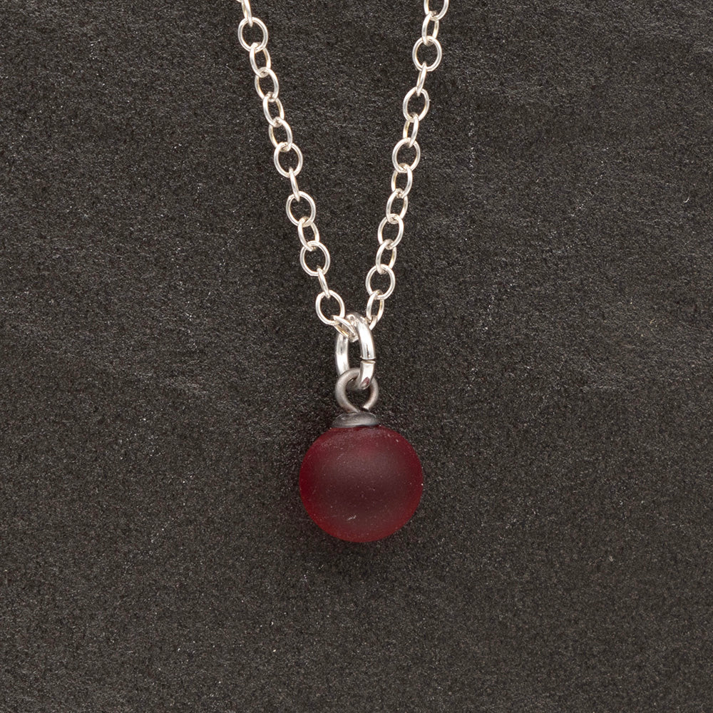 Close up of a  red frosted glass ball charm on a dark slate background. The charm is frosted. It hangs on a small silver jump ring. The necklace is completed with a silver chain.