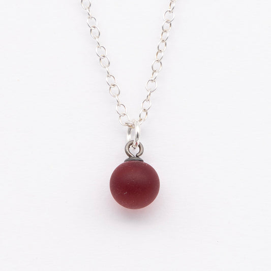 Close up of a red frosted glass ball charm on a plain white background. The charm is frosted. It hangs on a small silver jump ring. The necklace is completed with a silver chain.