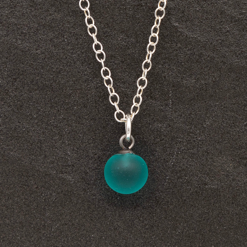 Close up of a teal frosted glass ball charm on a dark slate background. The charm is frosted. It hangs on a small silver jump ring. The necklace is completed with a silver chain.