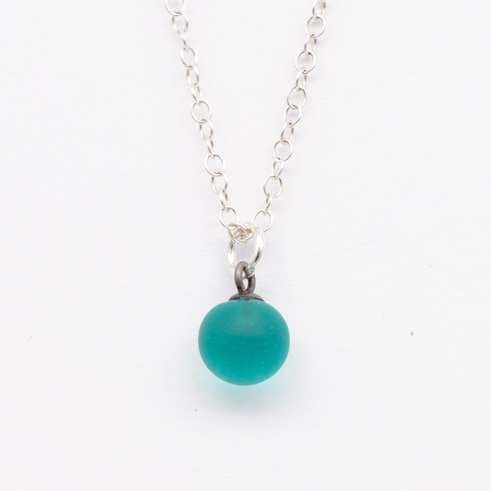 Close up of a teal frosted glass ball charm on a plain white background. The charm is frosted. It hangs on a small silver jump ring. The necklace is completed with a silver chain.