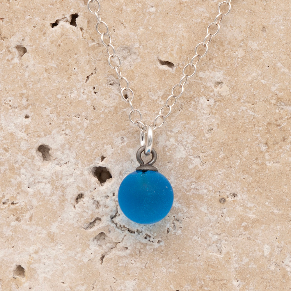 Close up of a turquoise frosted glass ball charm on a sandstone background. The charm is frosted. It hangs on a small silver jump ring. The necklace is completed with a silver chain.