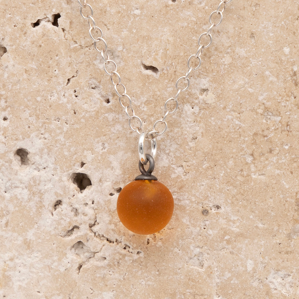 Close up of a yellow frosted glass ball charm on a sandstone background. The charm is frosted. It hangs on a small silver jump ring. The necklace is completed with a silver chain.