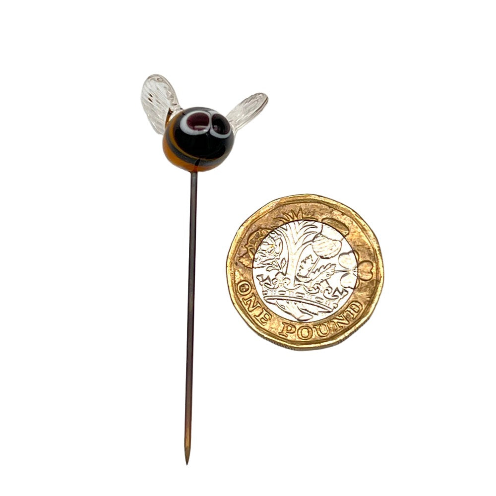 Size comparison of the handmade lampwork glass bee plant decoration placed next to a one-pound coin, highlighting its small size. The bee is on a stainless steel pin.