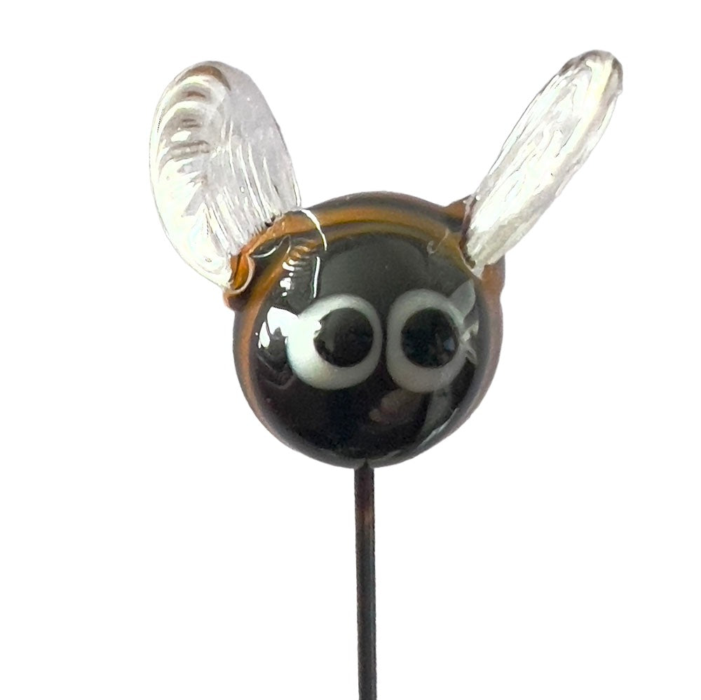 A close-up of a glass bee plant decoration viewed from the front. The bee has large, expressive eyes, translucent wings, and an amber and black striped body. It is mounted on a thin metal stake.