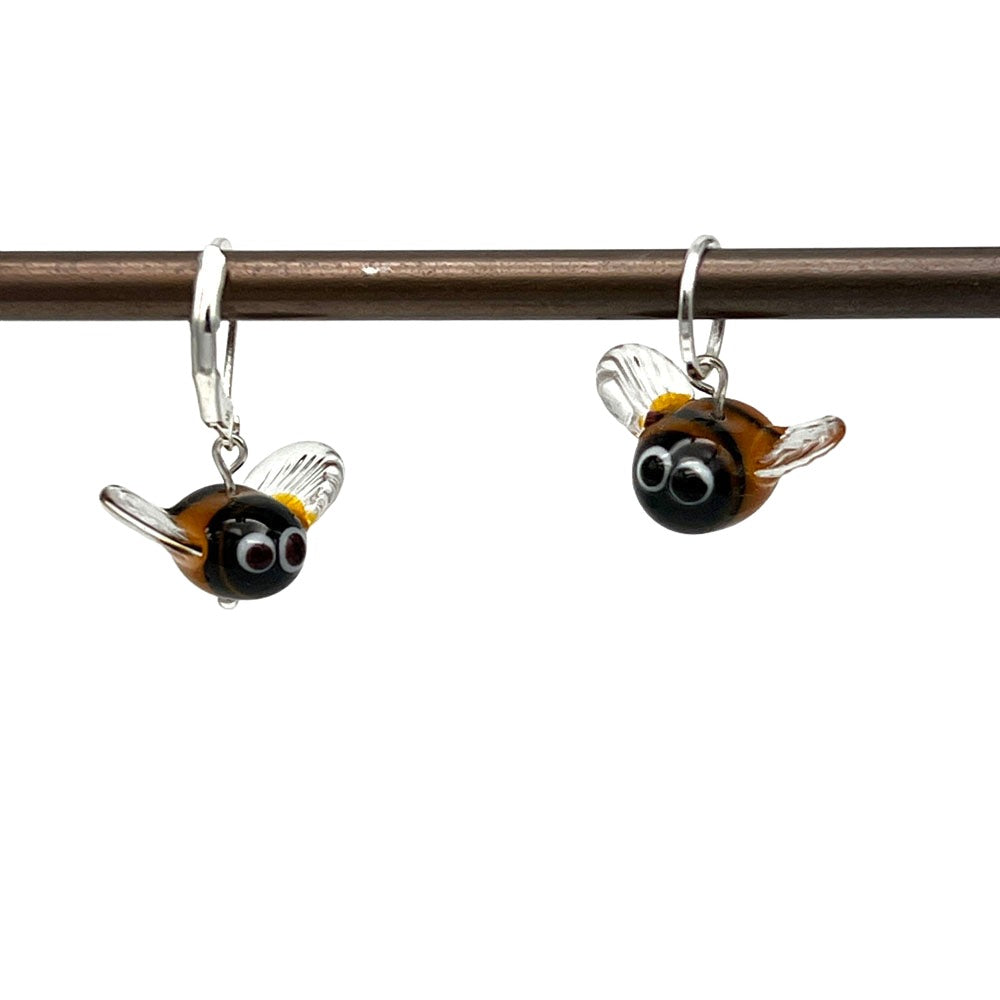 Bee Stitch Marker Hanging  : Image showing two handmade glass bee stitch markers hanging on a metal rod. Each bee has a black head with large white eyes, a yellow and black striped body, and clear wings.