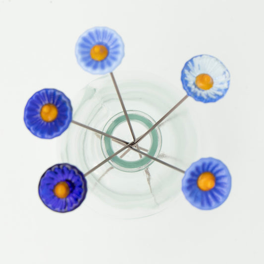 Five blue glass flowers shot from above. Flowers are different shades of blue and have yellow centres. The flowers sit on metal stakes.