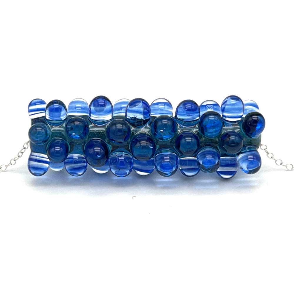 Close up of blue ripples bead necklace. The long tube bead is made of sparkly blue glass and is heavily decorated with dots made from layers of clear and transparent blue glass. The bead sits on a sterling silver chain.