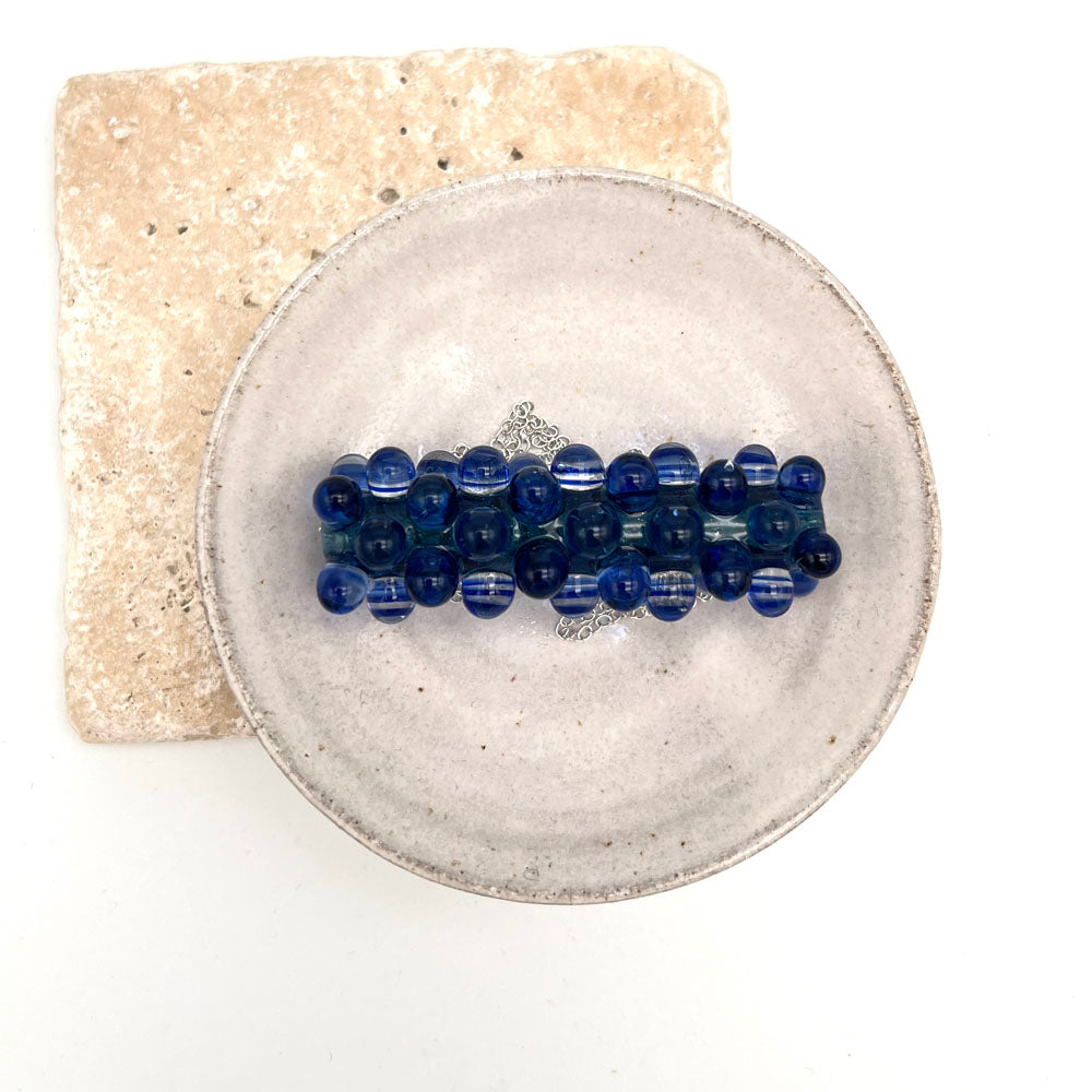 Blue ripples glass bead necklace sitting in a small white ceramic dish. Photographed from above.