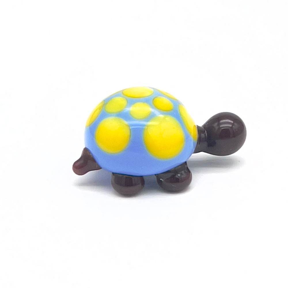 A small, glass tortoise with a blue shell decorated with yellow spots. The tortoise has a dark brown head and legs, displayed on a white background.