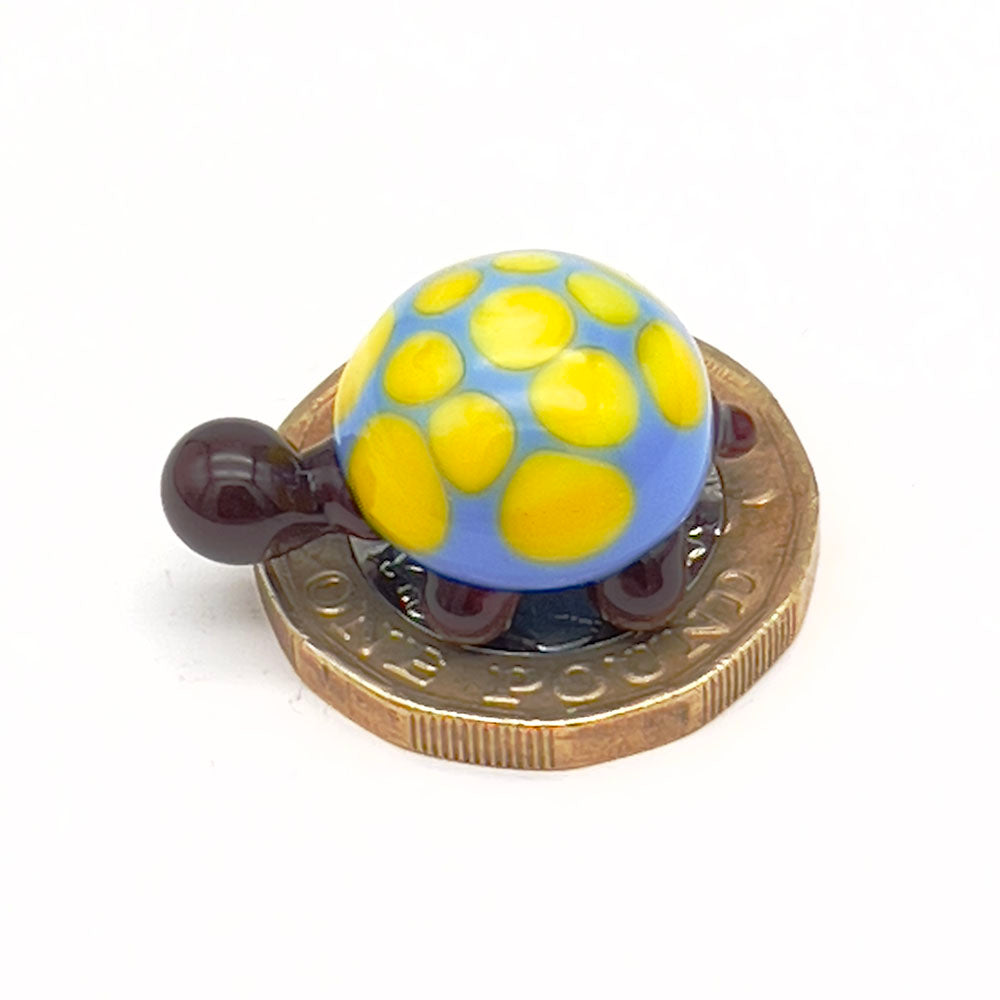 The miniature glass tortoise sitting on a one-pound coin, highlighting its small size. The tortoise has a blue shell with yellow spots, and dark brown head and legs.