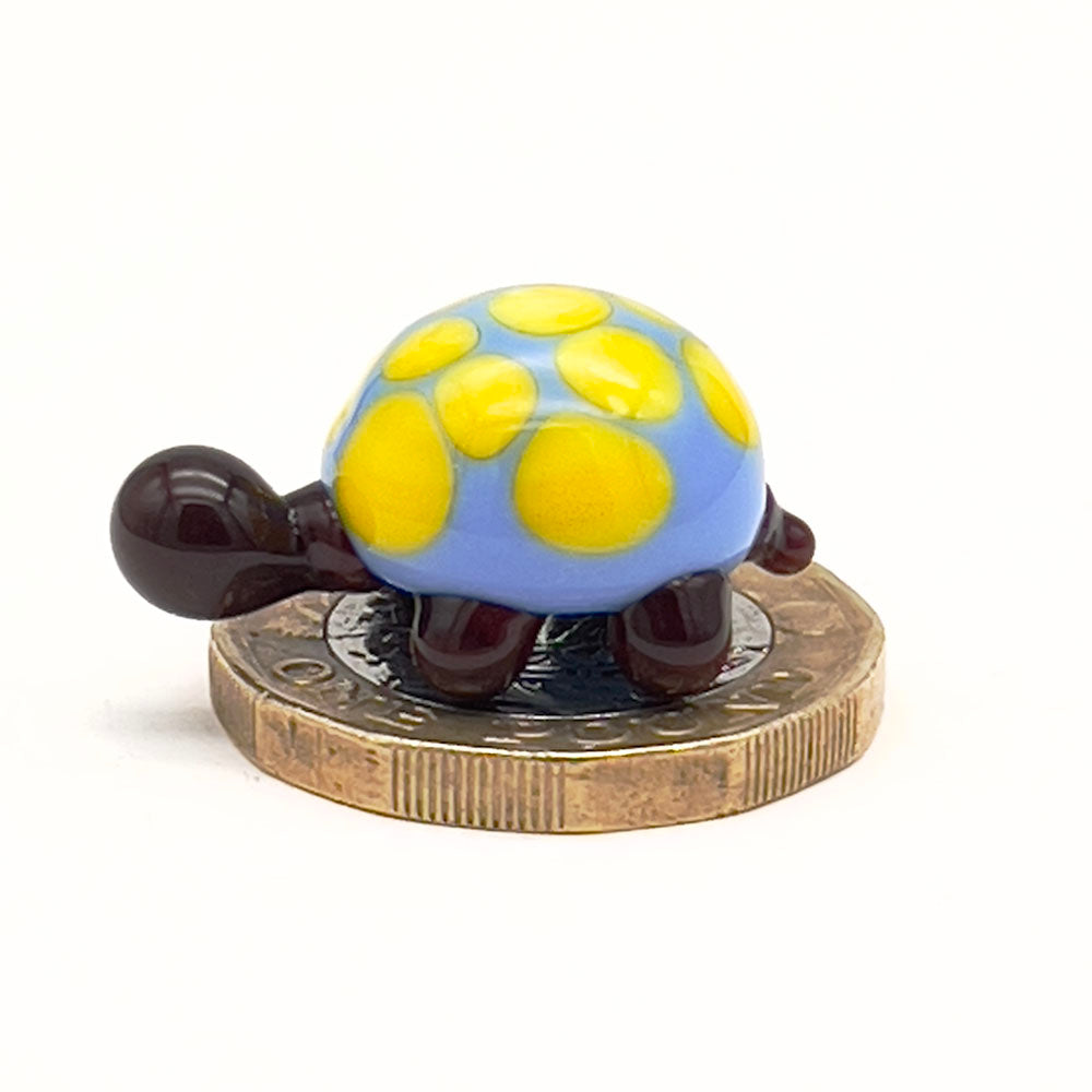 The glass tortoise on top of a one-pound coin, viewed from a different angle, emphasizing its small size. The tortoise features a blue shell with yellow spots and dark brown head and legs.