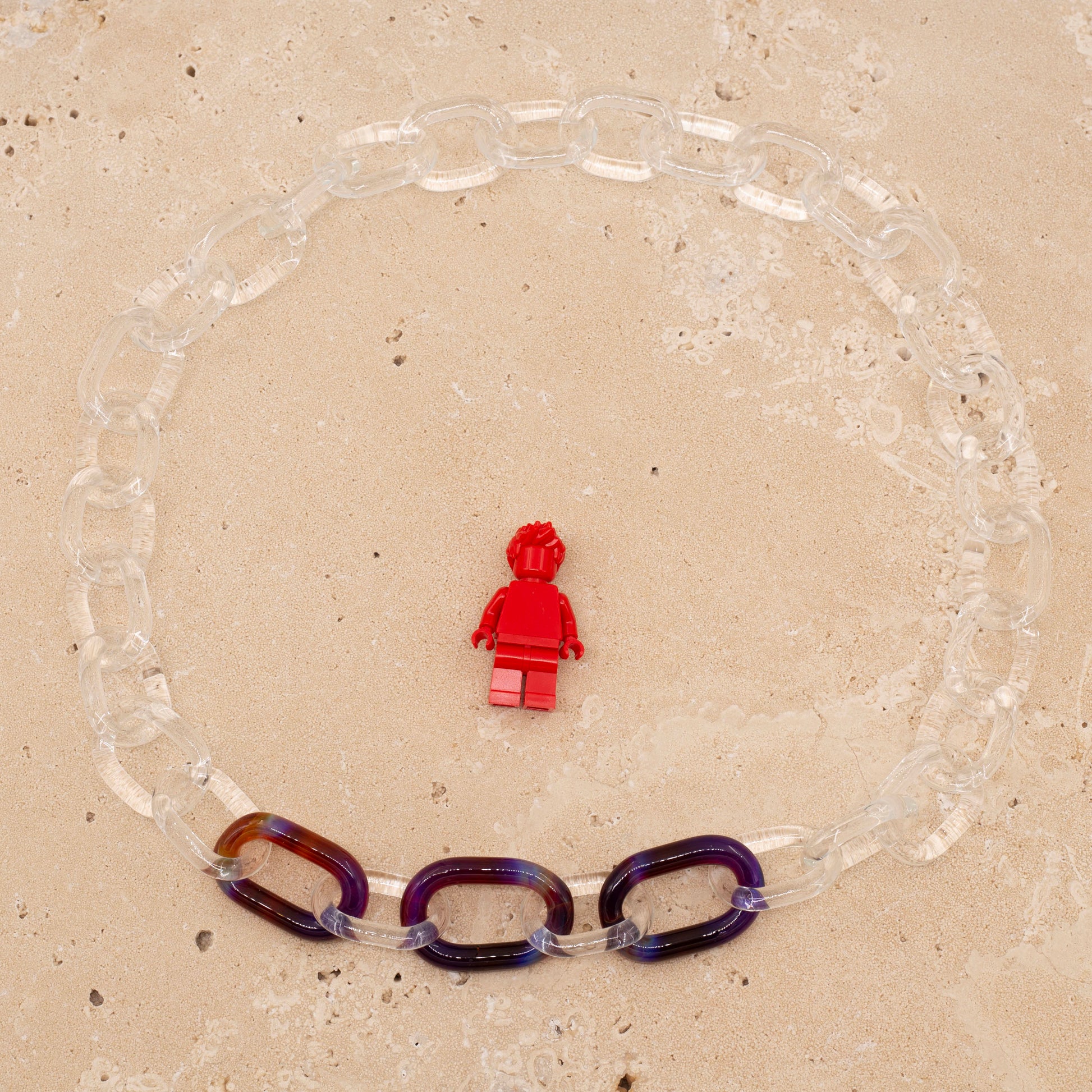 Overhead view of necklace made from clear glass links with 3 coloured links. Image includes a Lego figure for scale.