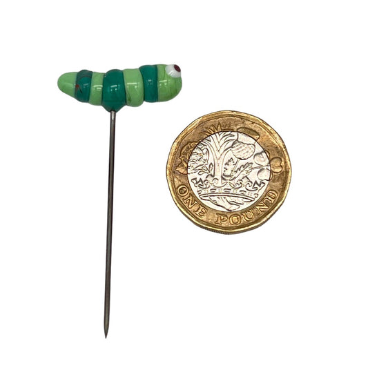 Handcrafted lampwork glass caterpillar pin next to a British one-pound coin for size comparison. The caterpillar is light green with dark green stripes.