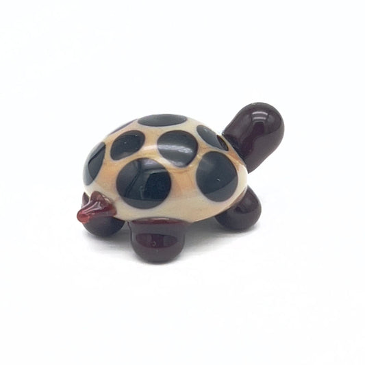 A small, glass tortoise with a beige shell decorated with dark brown spots. The tortoise has a dark brown head and legs, displayed on a white background.