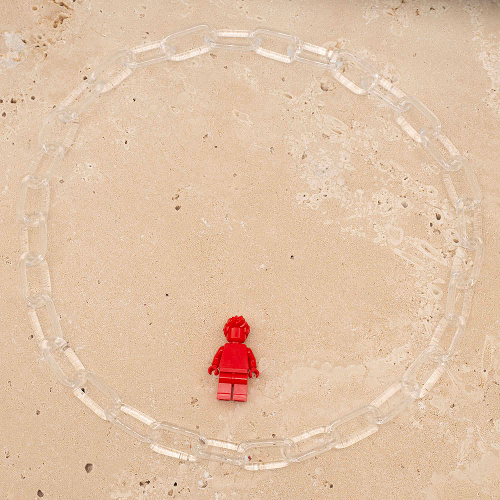 Clear glass chain link necklace photographed from above on sandstone tile. Shown with Lego figure for scale.