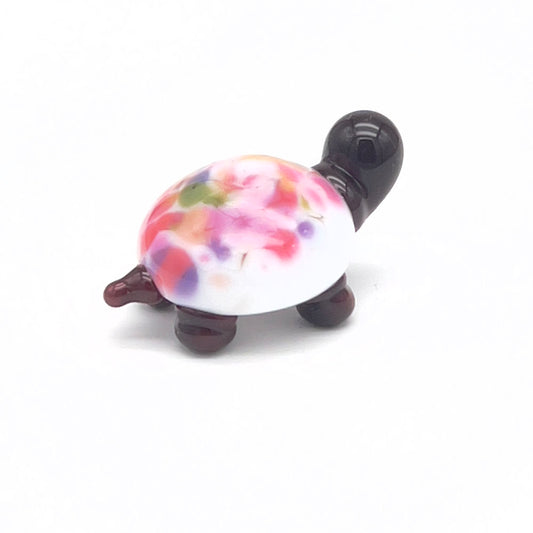 A small, glass tortoise with a white shell decorated with multicolored speckles in pink, purple, green, and orange. The tortoise has a dark brown head and legs, displayed on a white background.