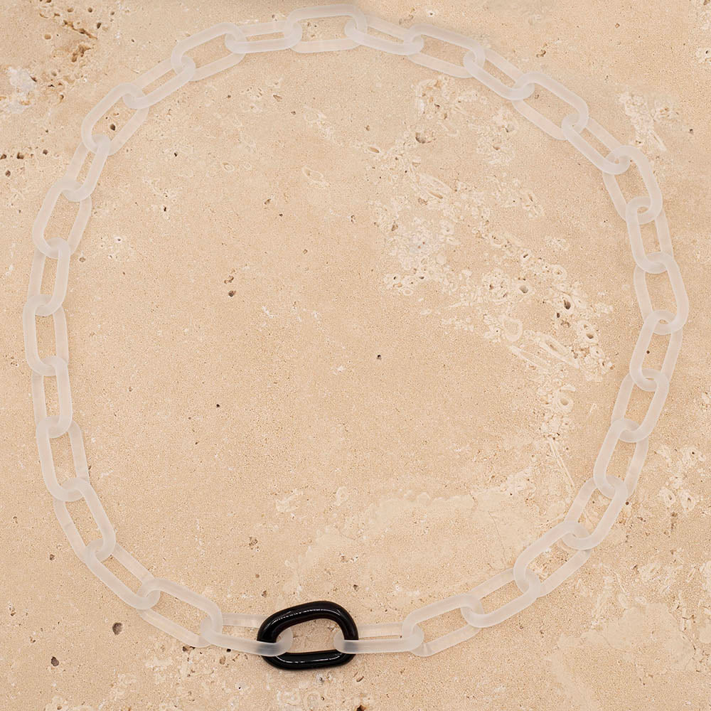 A clear frosted glass chain necklace with a single shiny glass link on a sandstone background