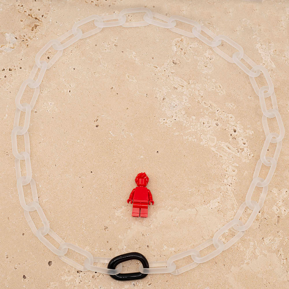 A clear frosted glass chain necklace with a single shiny glass link photographed from above with a Lego figure for scale