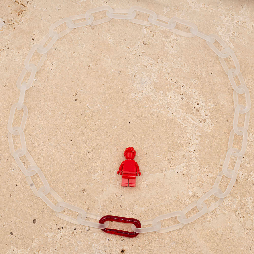 Clear frosted glass chain necklace with a single red link, photographed from above with Lego figure for scale.