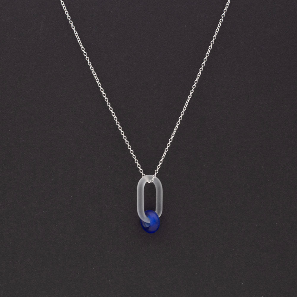 Dark background. Frosted clear glass link necklace. The link passes through a bead made form recycled blue sherry bottle glass, and the bead has a frosted finish. The link hangs from a sterling silver chain.