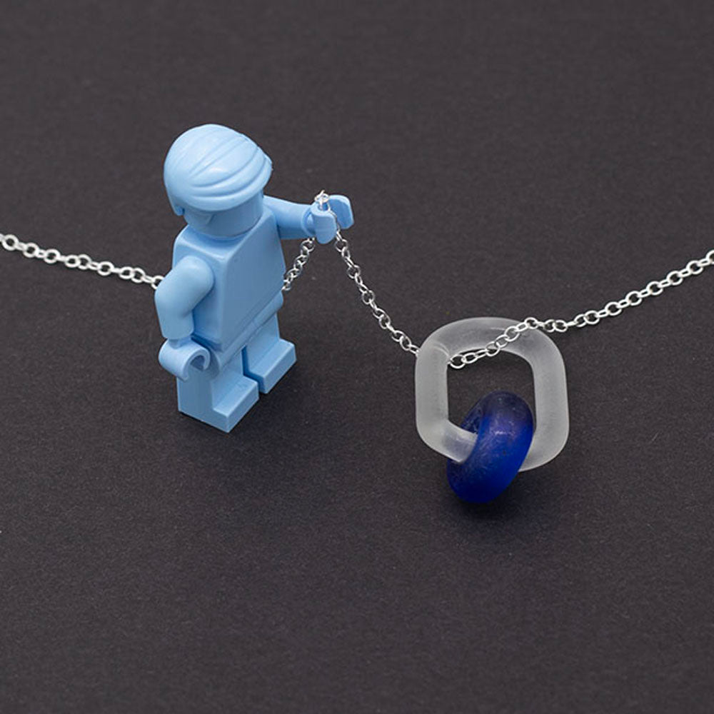 Necklace made with a clear glass link which passes though a bead made with blue glass from a sherry bottle. Bot bead and link have a frosted finish. The necklace is finished with a sterling silver chain. Photographed on a dark background with a pale blue Lego figure holding the chain