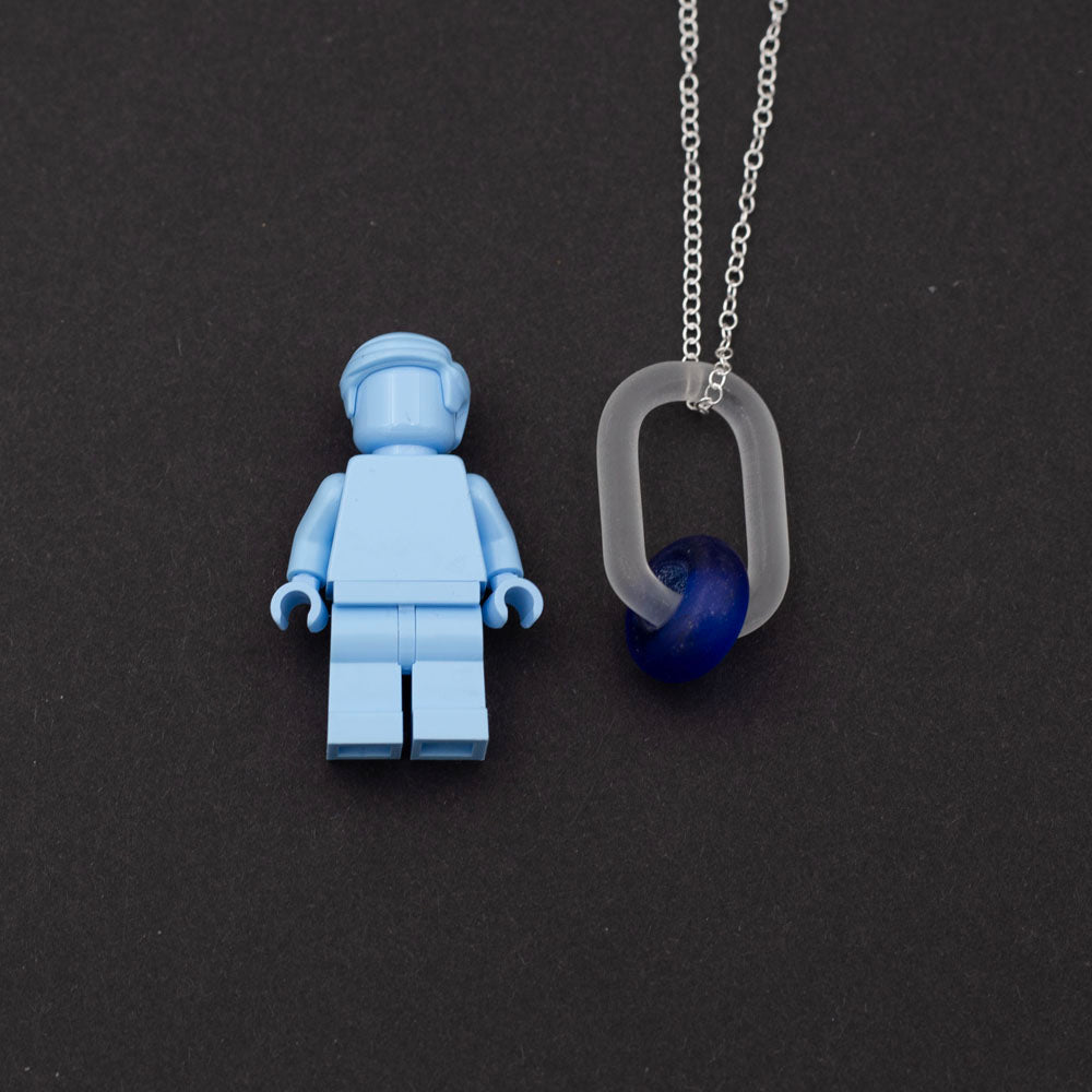 Lego figure with frosted clear glass link necklace. The link passes through a bead made form recycled blue sherry bottle glass, and the bead has a frosted finish. The link hangs from a sterling silver chain.