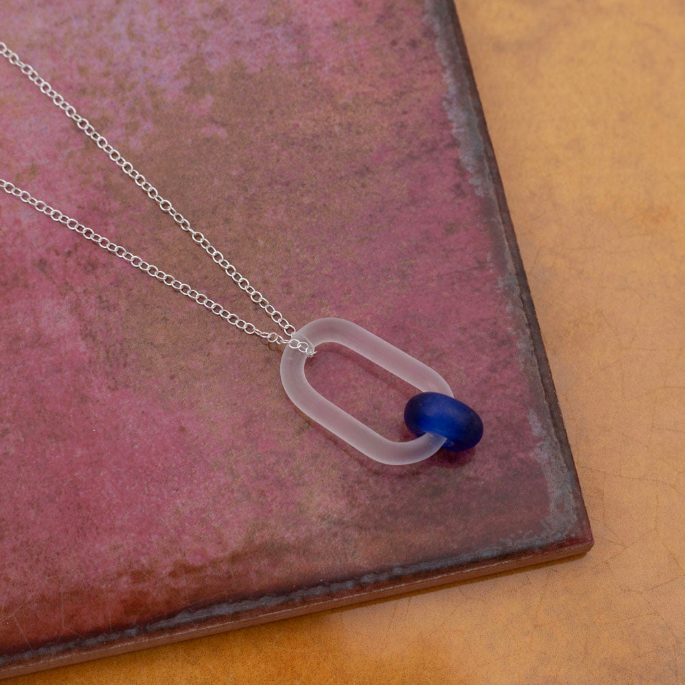 Necklace photographed on pink and yellow tiles. The link passes through a bead made form recycled blue sherry bottle glass, and the bead has a frosted finish. The link hangs from a sterling silver chain.