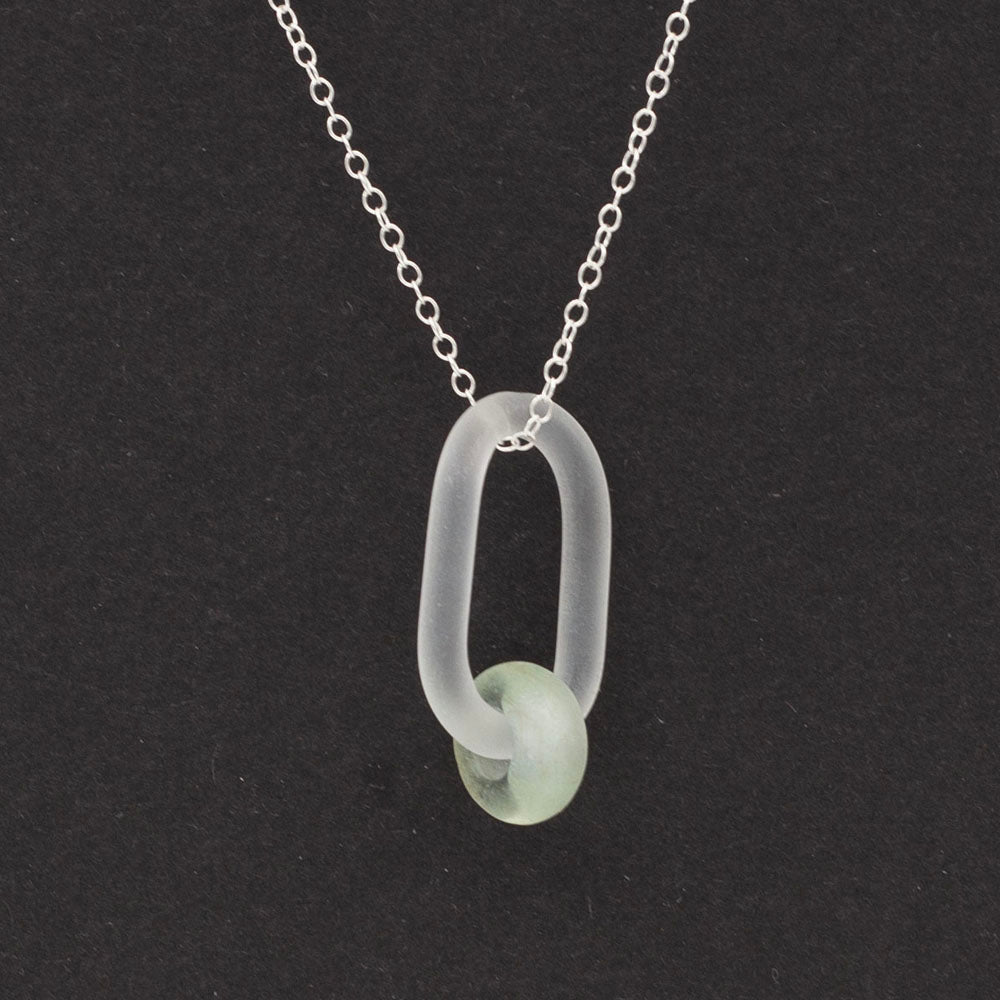 Close up view. Clear link which passes through a bead made from a recycled pale green wine bottle. Link and beads have a frosted finish. The link hangs from a sterling silver chain. Dark background.