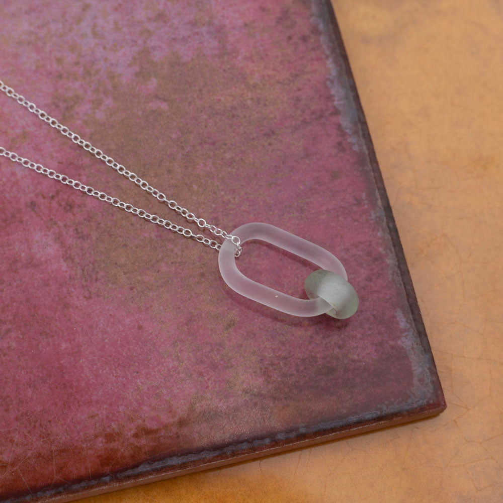 clear link which passes through a bead made from a recycled pale green wine bottle. Link and beads have a frosted finish. The link hangs from a sterling silver chain. Shown sitting on pink and yellow tiles.