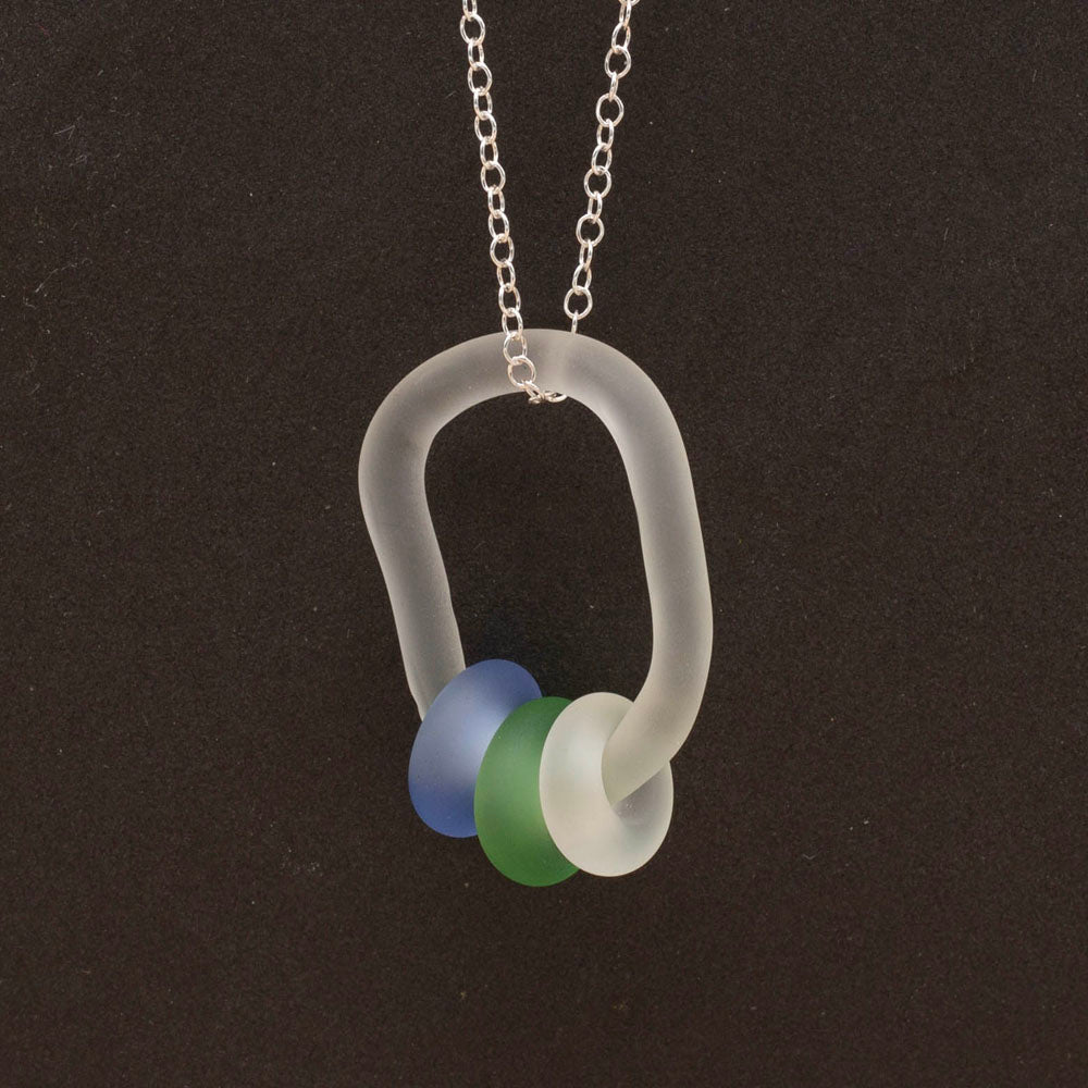 Close up of clear link which passes through beads in blue, green and clear glass. Link and beads have a frosted finish. The link hangs from a sterling silver chain. Dark background.
