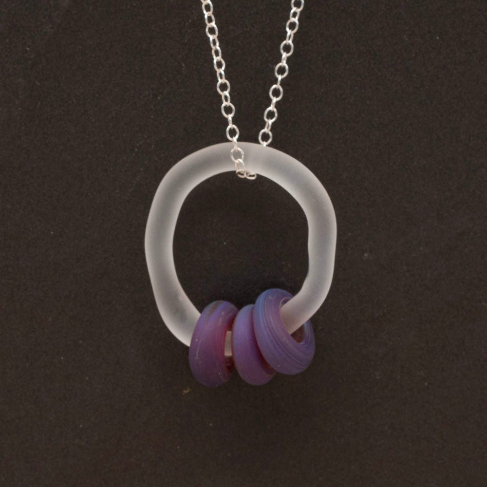 Close up of a necklace made with a glass link which passes through three purple beads. Both link and beads have a frosted finish. The necklace is completed with a sterling silver chain.