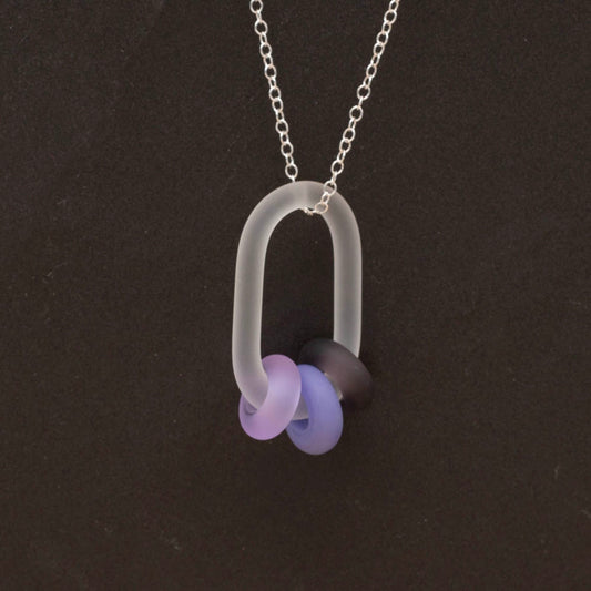 Close up of clear link which passes through beads in three shades of purple glass. Link and beads have a frosted finish. The link hangs from a sterling silver chain. Shown on dark background.