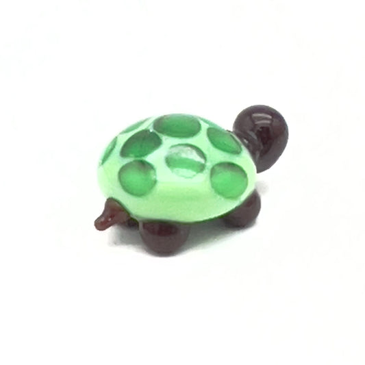 A small, glass tortoise with a light green shell decorated with dark green spots. The tortoise has a dark brown head and legs, displayed on a white background.