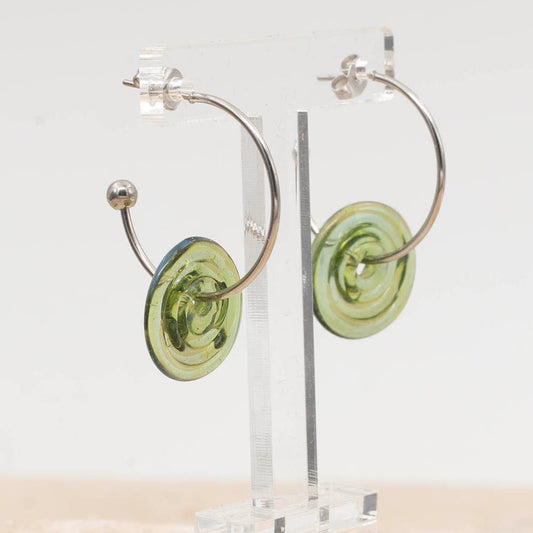 Stainless steel half hoop earrings with transparent green glass discs. The earrings have a ball at the end of the hoop and fasten with a butterfly scroll. The glass discs can move freely on the hoops. Shown on a clear earring stand.