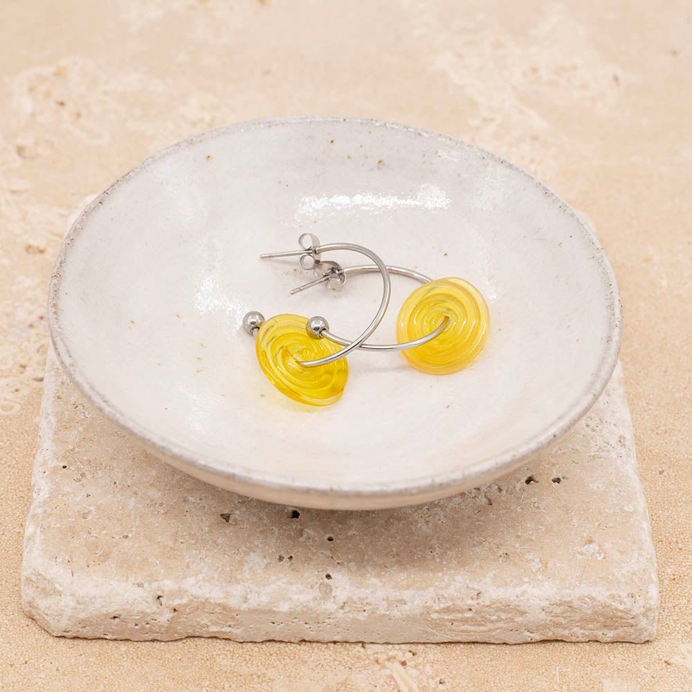 A pair of earrings made with sunshine yellow glass discs and stainless steel half hoop earrings. The earrings have a ball at one end and fasten with a butterfly scroll. Both earrings sit in a small cream pottery dish which sits on sandstone tiles.