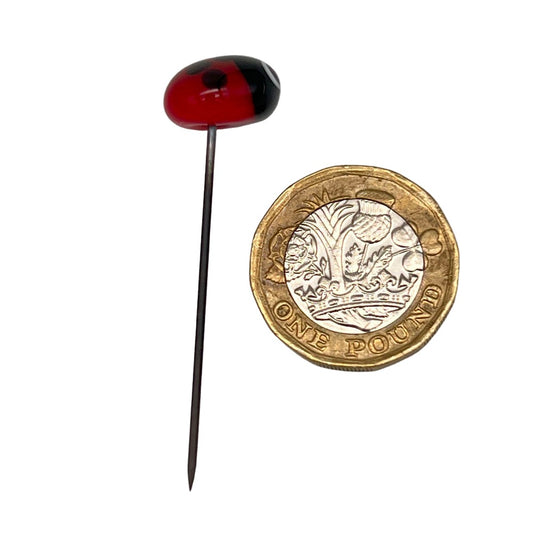 Handmade glass ladybird pin with a red body and black spots next to a one-pound coin for size comparison, on a stainless steel pin