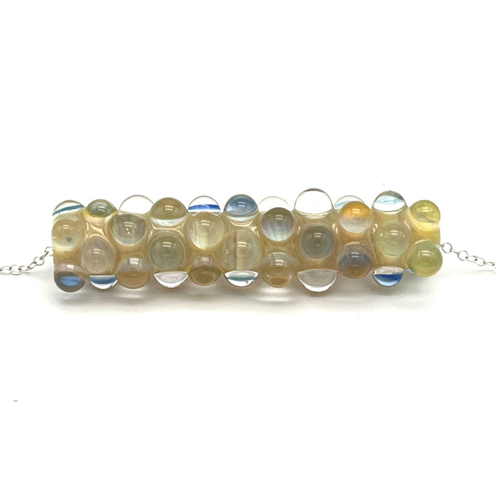 Close up of lemonade bead necklace. The bead is a long tube with core of many shades of yellow glass. The bead is heavily decorated with clear glass dots some of which include layers of transparent blue glass.