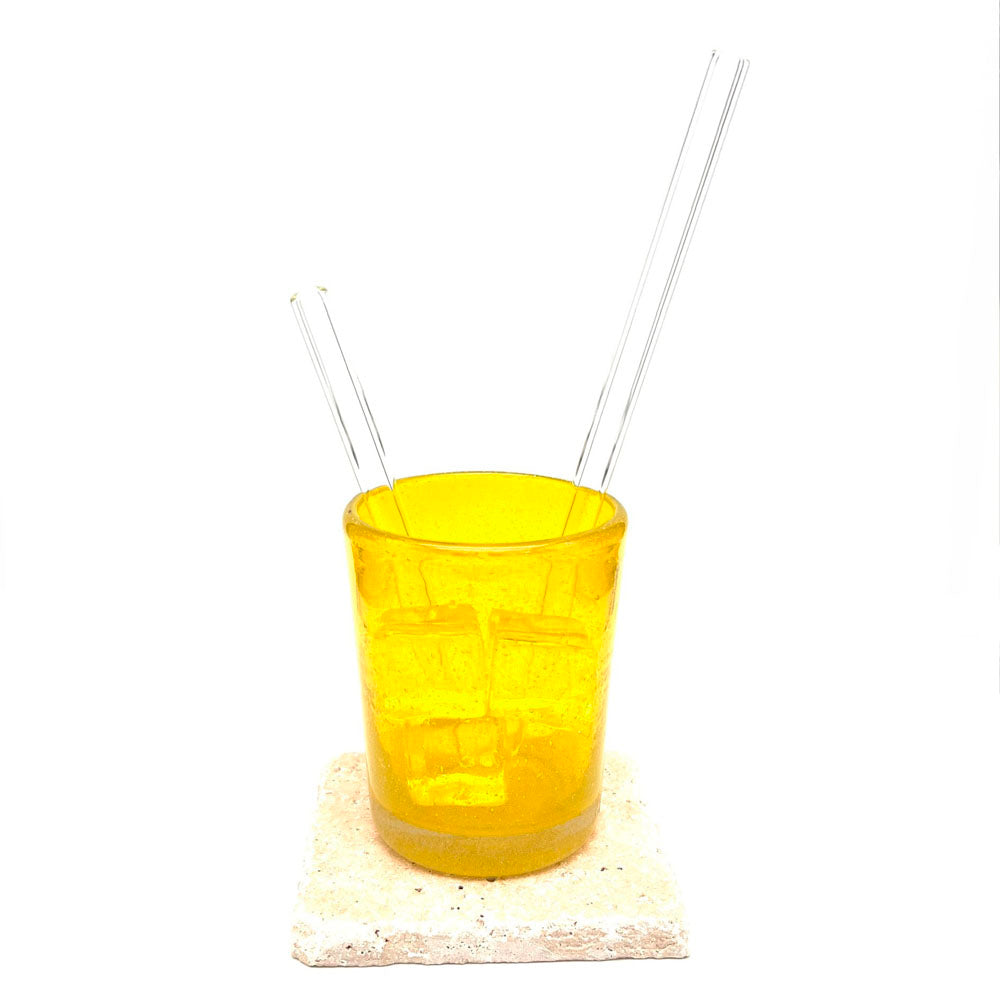 Long and short cler glass straws sitting in a yellow tumbler which is filled with ice. The tumbler sits on a sandstone tile.